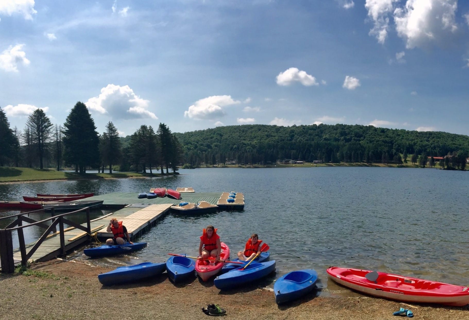 A group of people sit in kayaks on a dock overlooking a tranquil lake in western New York, enjoying their camping experience among cozy cabins.
