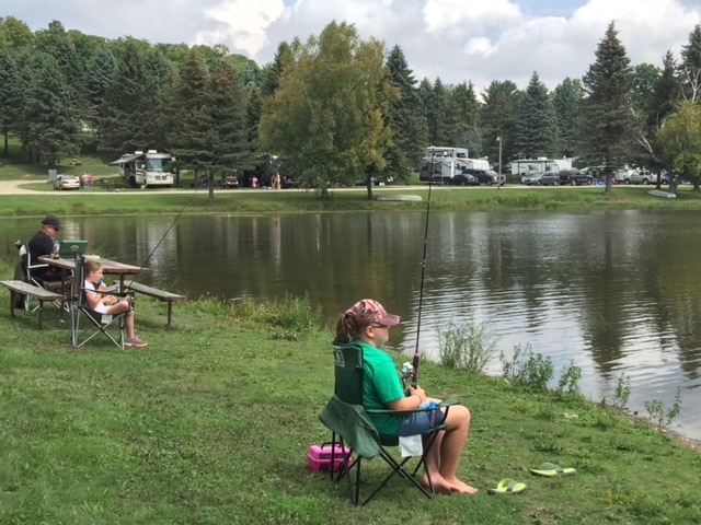 A family sits on chairs near a pond and fishes during their camping trip in Western New York.