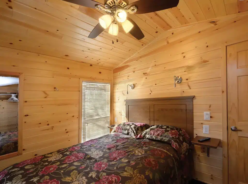 Main bedroom inside a park model cabin, with a queen bed, vaulted pine ceiling and wall-mounted reading lights