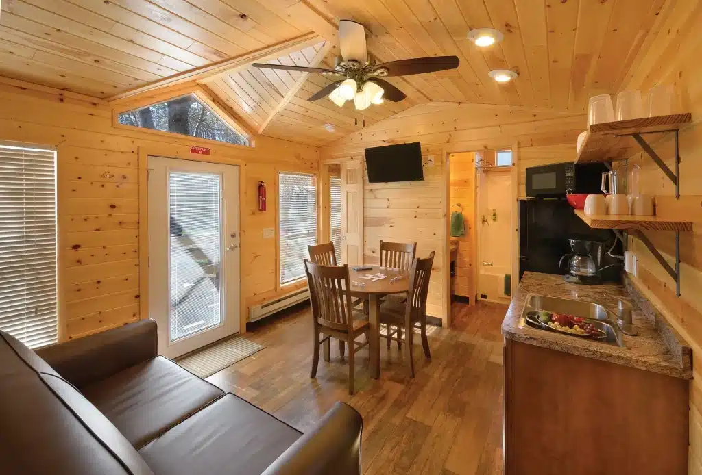 Open living, dining and kitchen area inside a park model cabin, with a vaulted pine ceiling, ceiling fan and wood floors