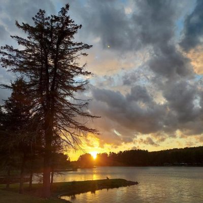 The sun is setting over Rainbow Lake in western New York with trees in the background.